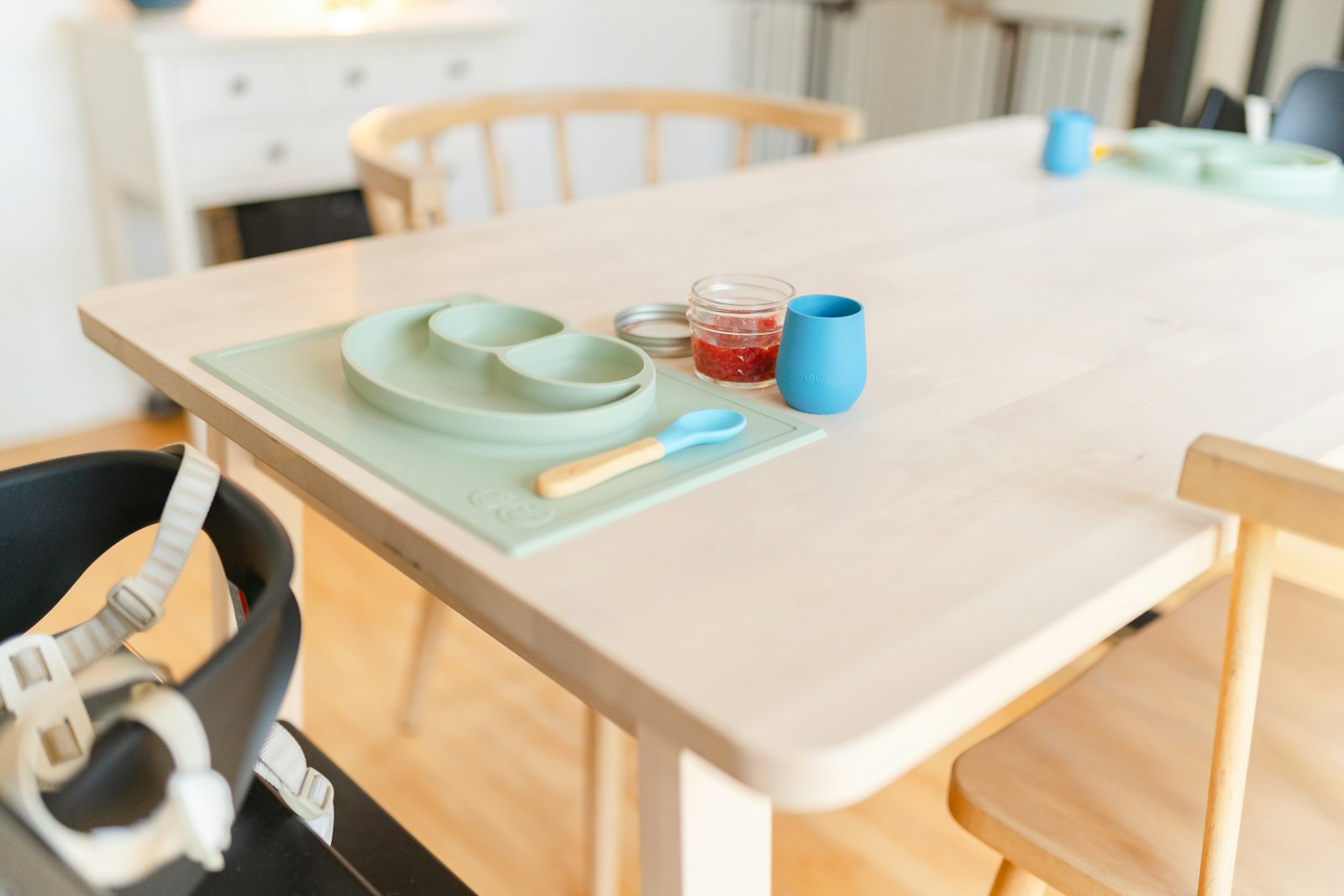 A wood dining table set with a sage silicone divided baby plate, a small wood-handled baby spoon, a glass jar of red puree, and a blue silicone open cup, with a high chair seat in the foreground.