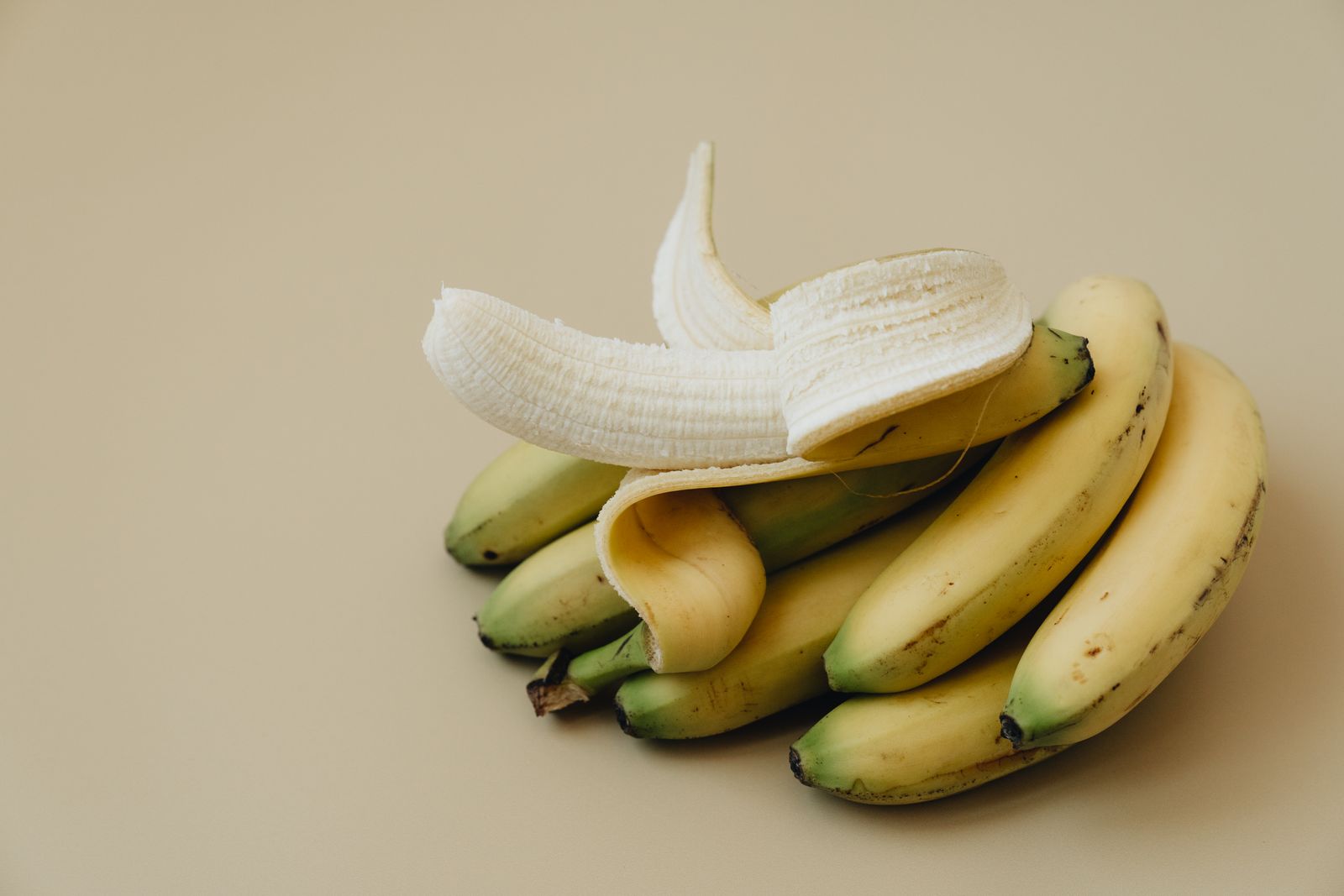 A ripe banana peeled open and resting on a small bunch of yellow bananas against a warm cream background.
