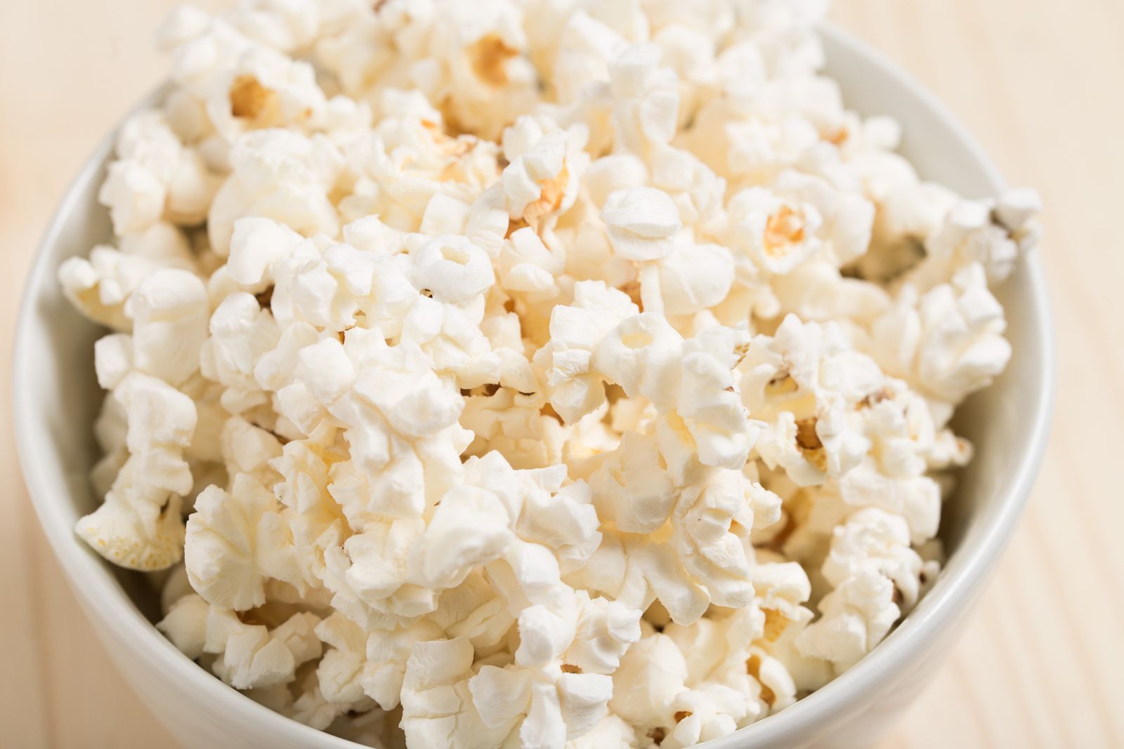 A close-up of freshly popped white popcorn filling a white ceramic bowl on a light wooden surface.