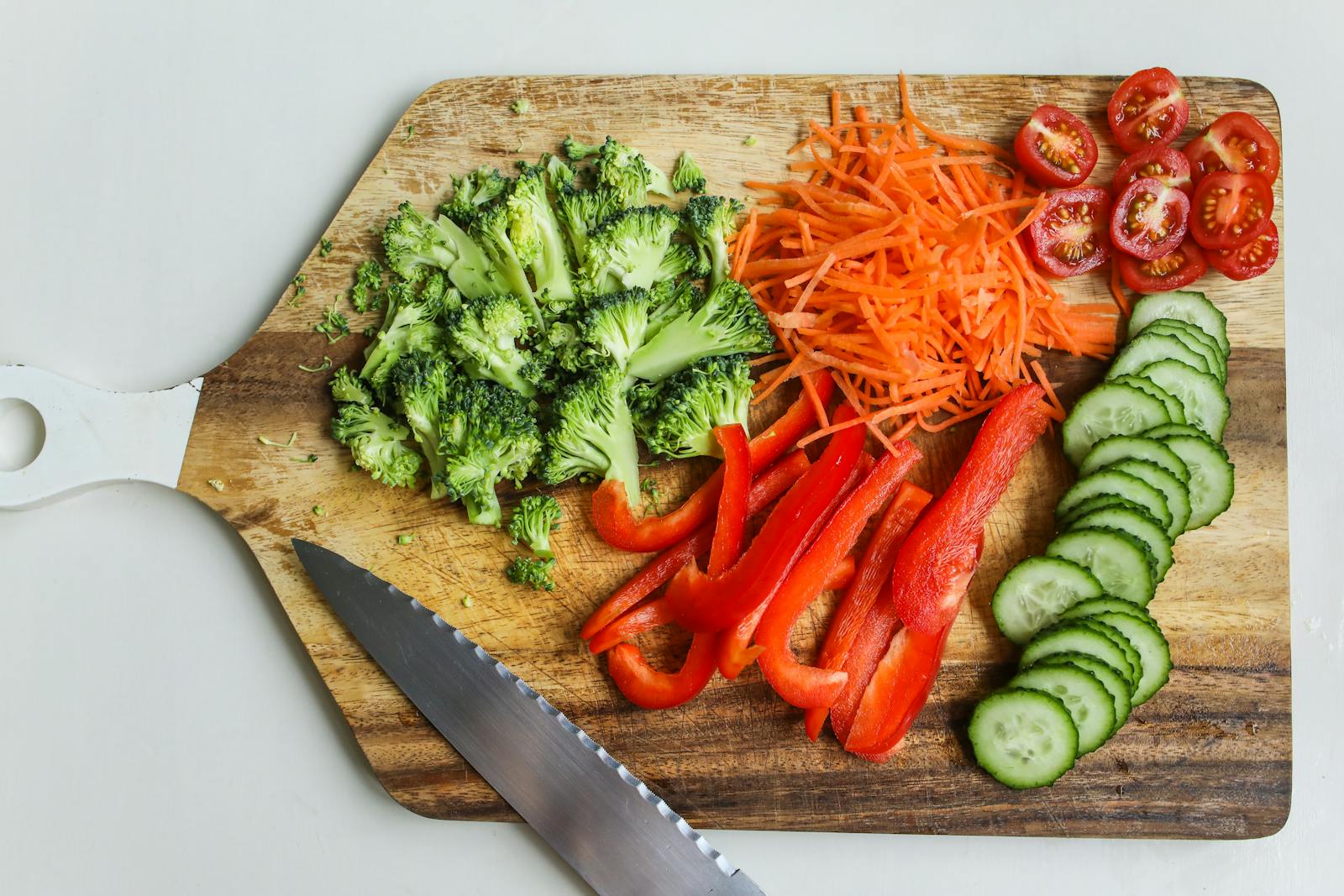 Overhead view of a wooden cutting board with halved cherry tomatoes, julienned carrots, broccoli florets, red pepper sticks, and sliced cucumber rounds beside a knife.