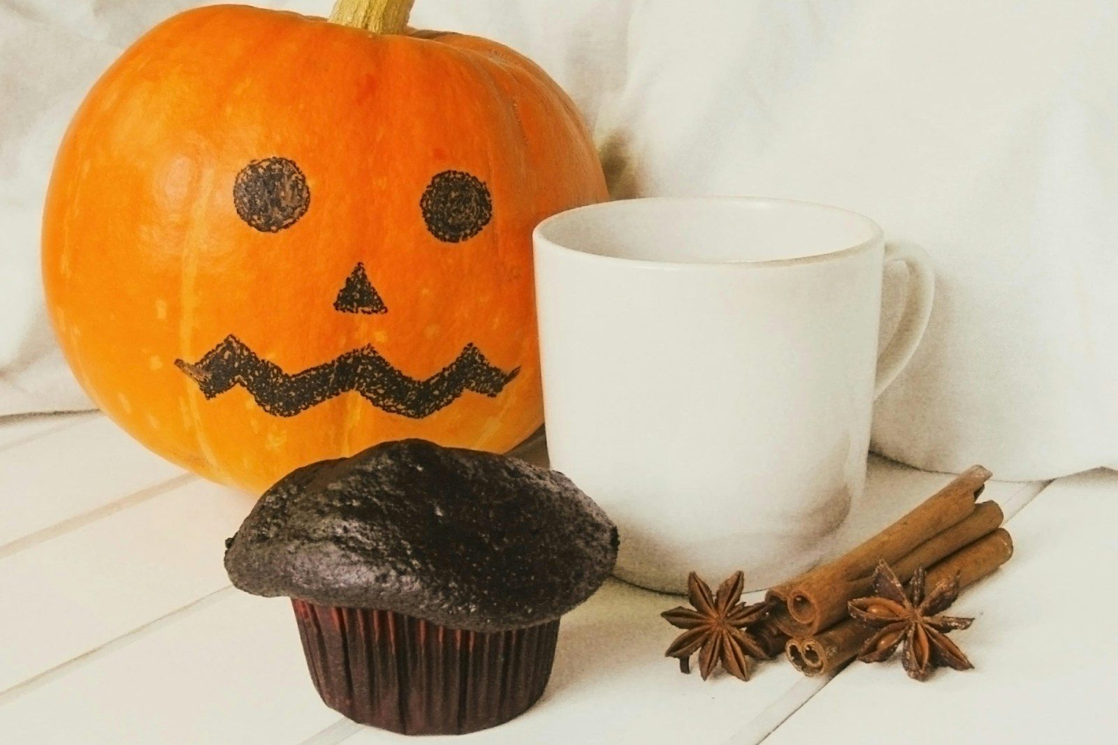 A jack-o'-lantern pumpkin beside a chocolate muffin, a white mug, cinnamon sticks and star anise on a white wooden table.