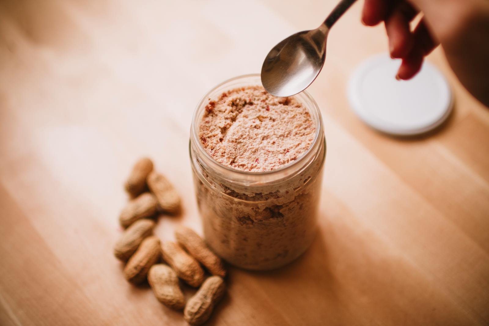 A hand reaching with a spoon to scoop natural peanut butter from a small glass jar on a warm wood table, with whole peanuts in shells beside it.