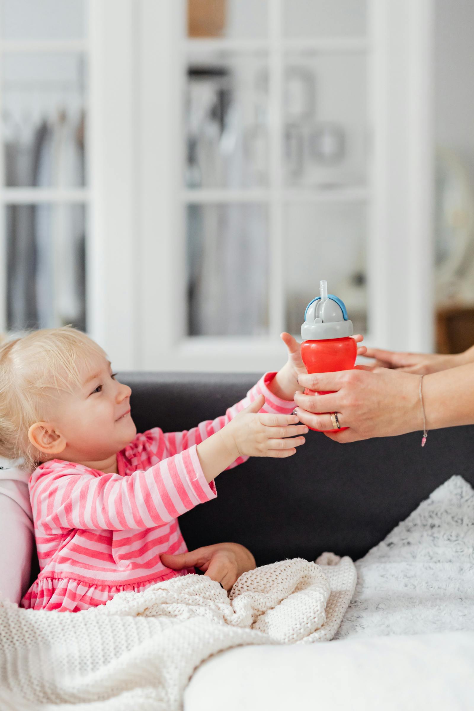 A young toddler in pink-striped pajamas reaches up from a couch toward a red sippy cup held by an adult's hand.
