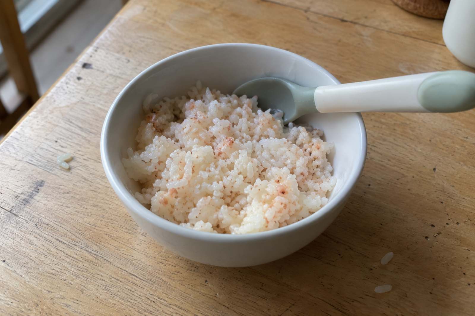 A small white bowl of soft rice mixed with finely minced cooked shrimp, with a baby spoon resting on the rim.