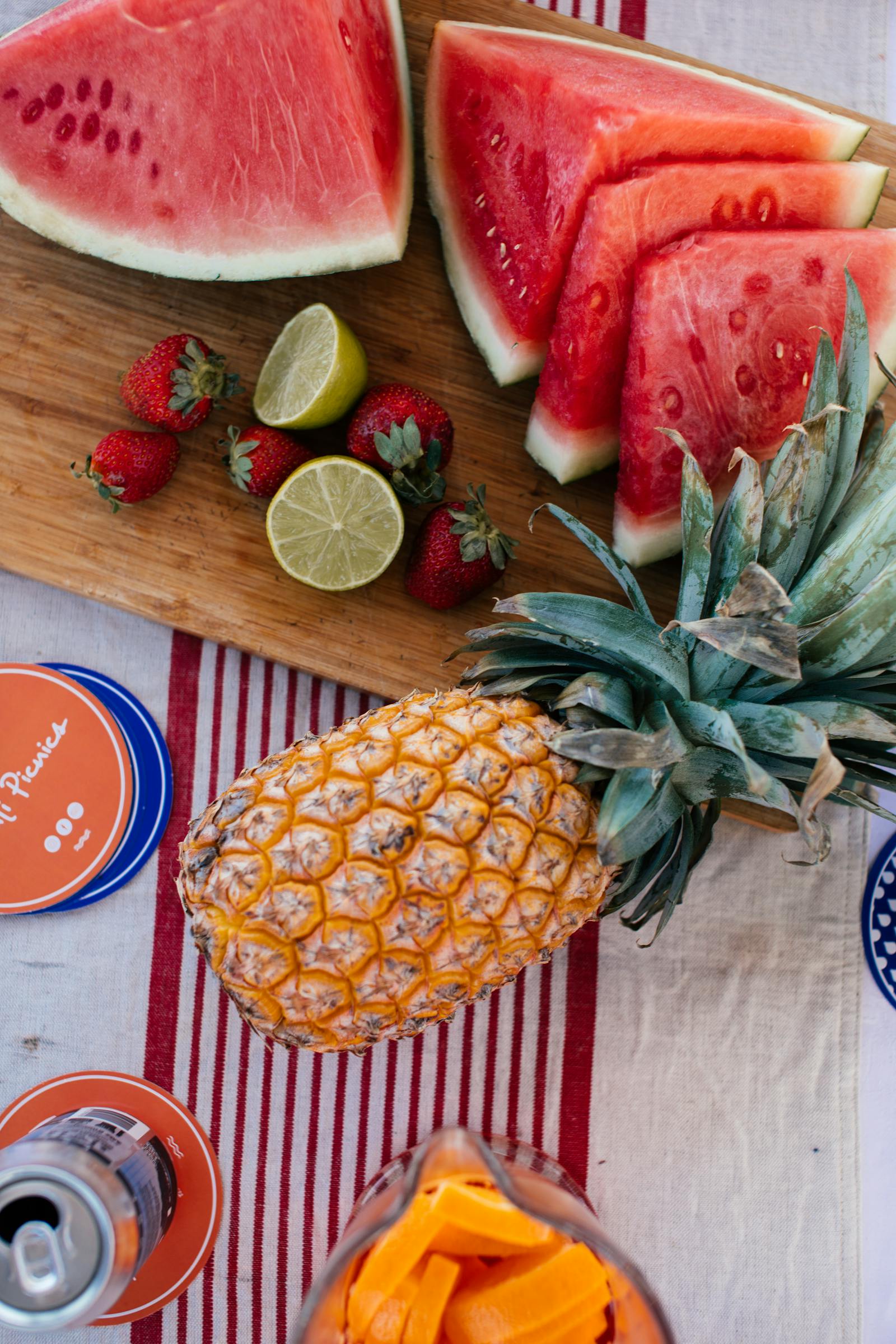 An overhead shot of a wooden board with watermelon wedges and slices, strawberries, halved limes, a whole pineapple, and a bowl of cut mango on a red-striped tablecloth.