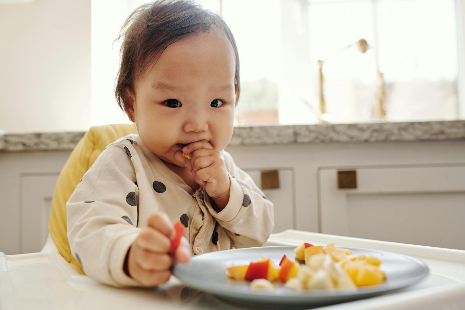 A baby in polka-dot pajamas sitting in a high chair, reaching for a plate of diced peach, banana, and apple while bringing a piece to the mouth.