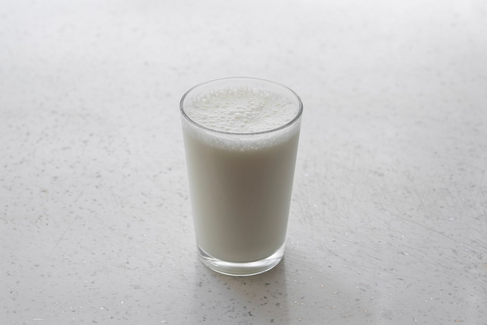 A short glass filled with cold milk, topped with a light layer of foam, on a pale speckled surface.